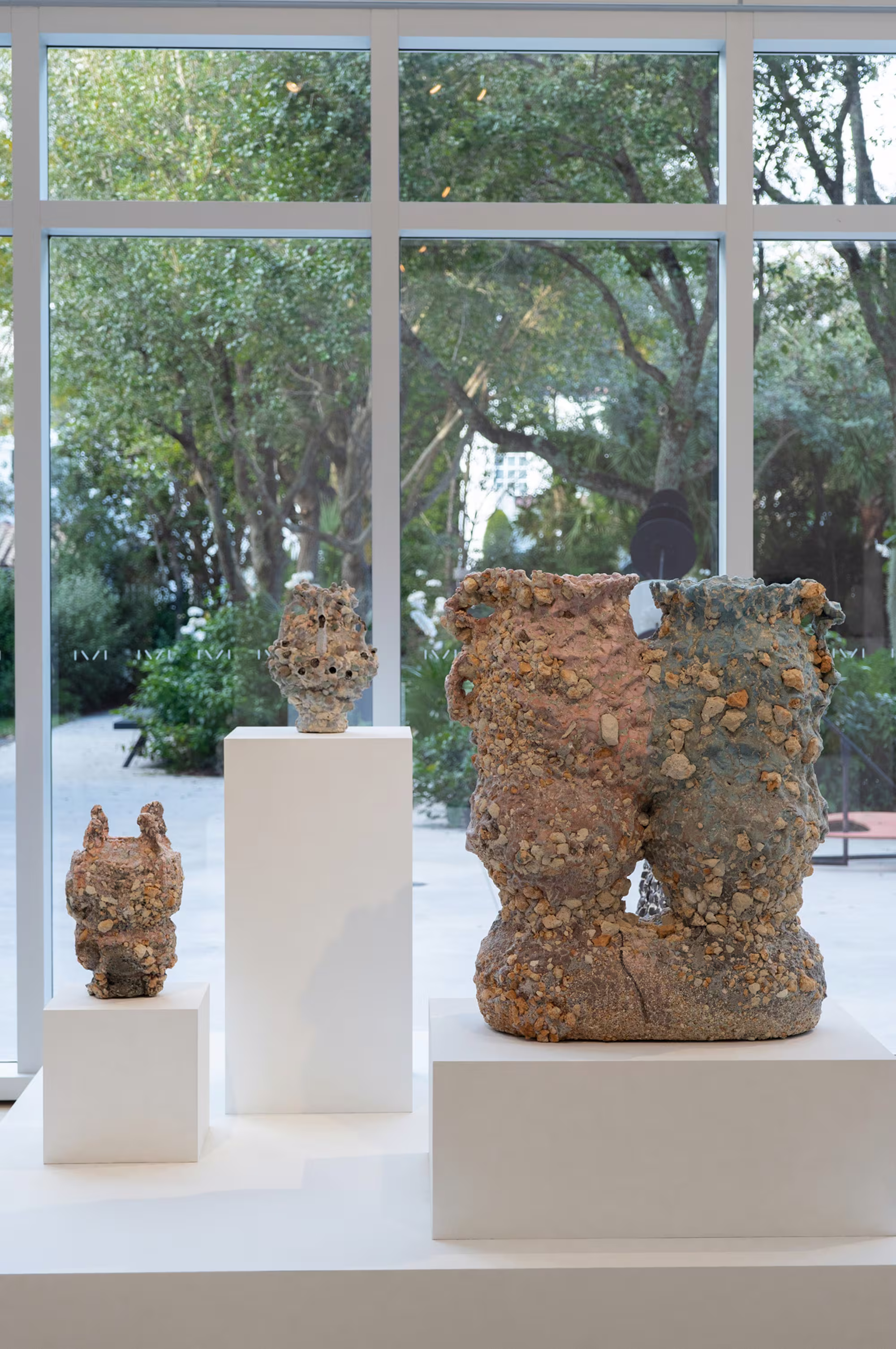 Three textured ceramic sculptures on white pedestals in front of large windows overlooking a lush garden at ICA Miami.