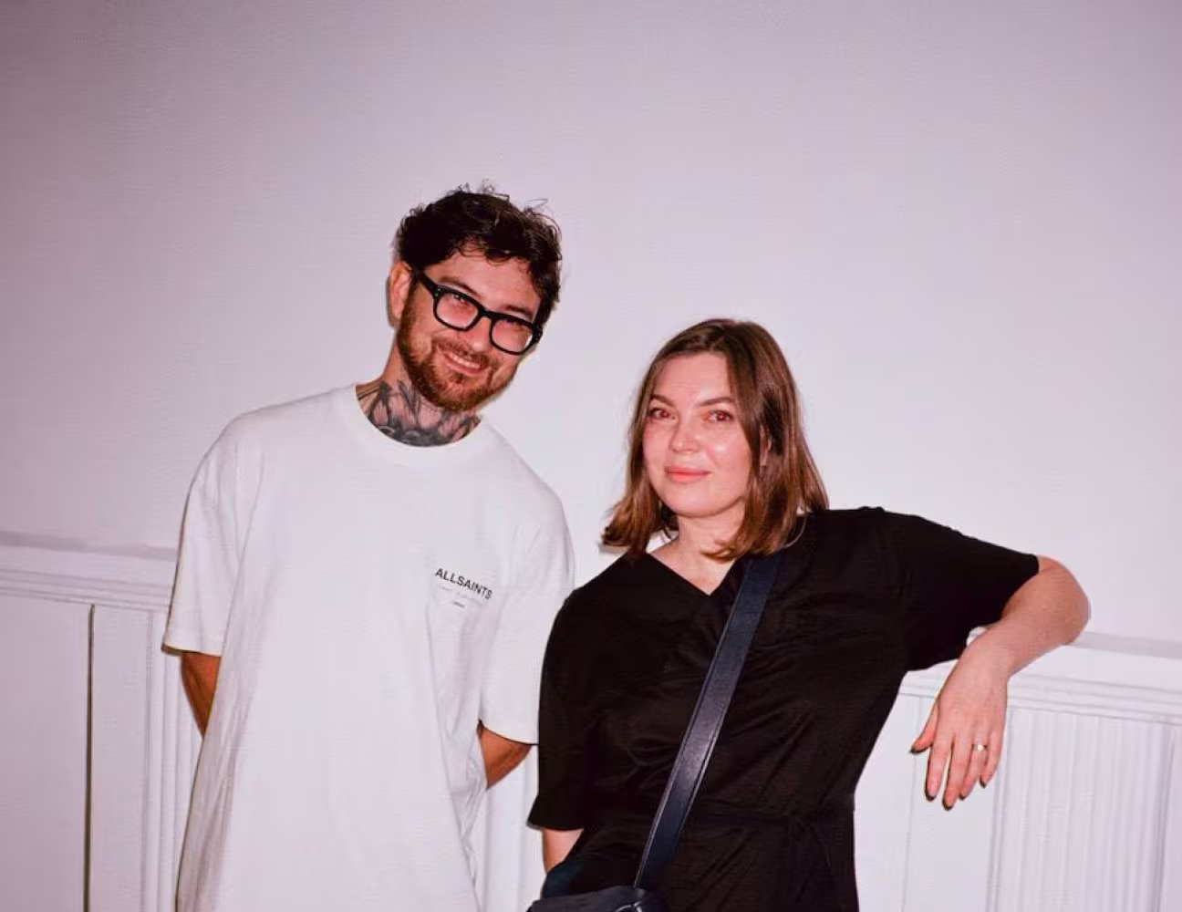 Two people smiling, one with glasses and tattoos wearing a white T-shirt, the other with shoulder-length hair in a black outfit. Minimalist white background.